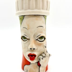 May include: A ceramic art piece in the shape of a pill bottle, featuring a sculpted face with green eyes, red lips, and a hand giving the middle finger. The bottle has a white cap and red and green accents.
