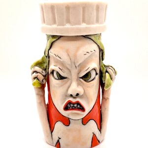 May include: A ceramic figurine of a person with an angry expression. The figurine is wearing a white chef's hat and has green hair. The figurine is painted in shades of red, white, and green.