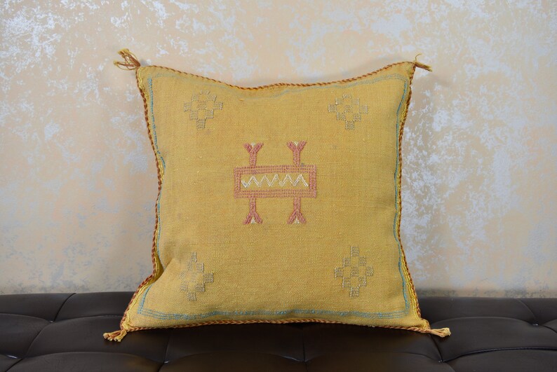 May include: A yellow throw pillow with a geometric pattern in pink and white. The pillow has tassels on the corners.