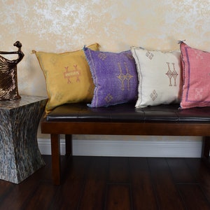 May include: Four decorative throw pillows with geometric patterns in shades of yellow, purple, white, and pink. The pillows are arranged on a brown leather bench with a wooden frame.