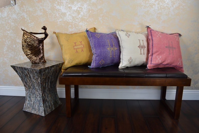 May include: Four decorative throw pillows with geometric patterns in shades of yellow, purple, white, and pink. The pillows are arranged on a brown leather bench with a wooden frame.