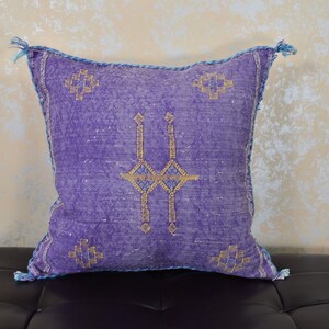 May include: Purple throw pillow with a geometric pattern in gold and blue thread. The pillow has a fringe border.