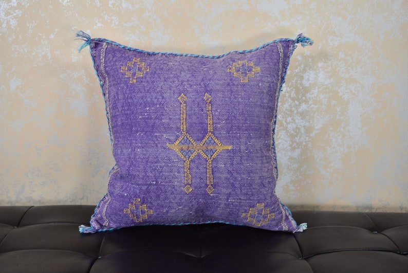 May include: Purple throw pillow with a geometric pattern in gold and blue thread. The pillow has a fringe border.