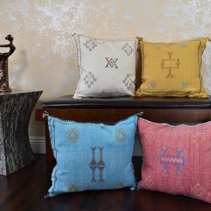 May include: Four decorative throw pillows in cream, yellow, light blue, and red. Each square pillow features a geometric design in contrasting colors. The pillows are placed on a dark brown bench. The background includes a decorative sculpture and a textured wall.