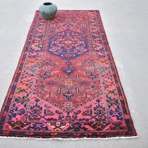 May include: A rectangular, hand-knotted rug with a central medallion design. The rug features a vibrant color palette of pink, red, blue, and touches of green and orange. A decorative, green vase sits at the top.