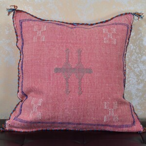 May include: A pink throw pillow with a traditional Moroccan design. The pillow has a blue and brown trim and features a geometric pattern in the center.