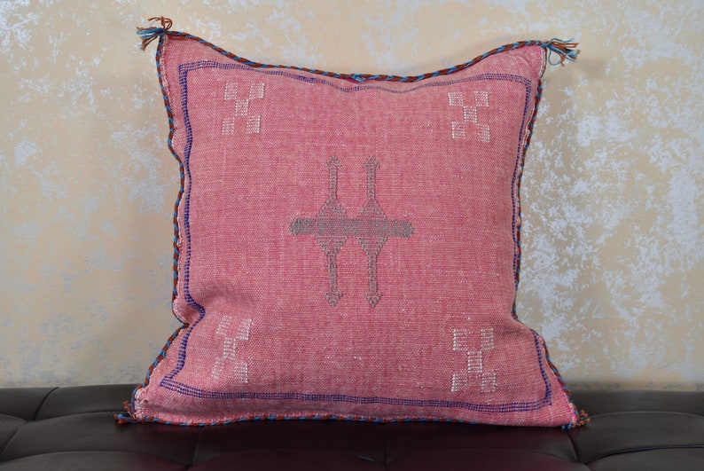 May include: A pink throw pillow with a traditional Moroccan design. The pillow has a blue and brown trim and features a geometric pattern in the center.