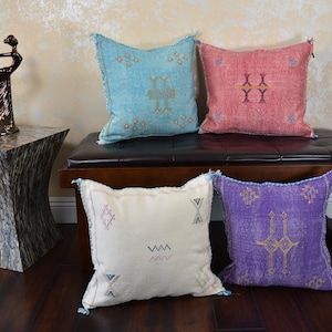 May include: Four square decorative pillows in shades of blue, red, white, and purple with embroidered geometric patterns. The pillows are arranged on a dark brown bench. A bronze sculpture and a decorative side table are also in the image.