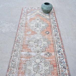 May include: A distressed, vintage-style runner rug with a central geometric pattern in cream, orange, and brown. The rug has a faded appearance and a decorative border. A green vase sits on the rug.