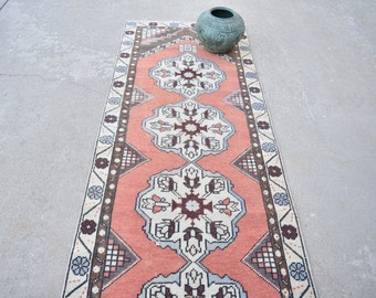 May include: A rectangular, coral-colored runner rug with a central floral pattern in white, gray, and burgundy. The rug has a border with geometric and floral designs in complementary colors. A teal vase sits on the rug.