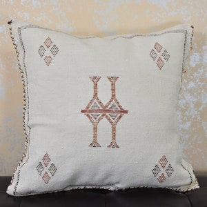 May include: White throw pillow with a geometric pattern in brown and red thread embroidery. The pillow has a diamond shape in each corner and a larger diamond shape in the center.
