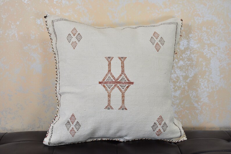 May include: White throw pillow with a geometric pattern in brown and red thread embroidery. The pillow has a diamond shape in each corner and a larger diamond shape in the center.