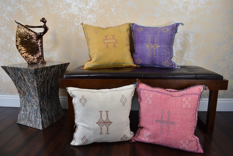 May include: Four decorative throw pillows with geometric patterns in shades of yellow, purple, white, and pink. The pillows are arranged on a brown wooden bench with a black leather top.