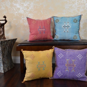 May include: Four square decorative throw pillows in shades of red, blue, yellow, and purple with embroidered geometric patterns. The pillows are arranged on a dark brown bench. Tassels adorn the corners of the pillows.
