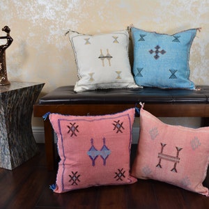 May include: Four decorative throw pillows with geometric patterns in cream, light blue, and pink hues. The pillows are arranged on a dark brown bench. A bronze sculpture and a wooden side table are also in the image.