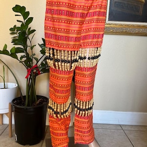 May include: Orange and brown patterned trousers with black and wooden bead fringe. The trousers have a wide waistband and are made of a lightweight fabric.