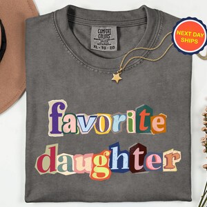 May include: A charcoal grey Comfort Colors t-shirt with the words "favorite daughter" in colourful, cut-out style letters. A gold star pendant necklace and a "Next Day Ships" sticker are also visible.