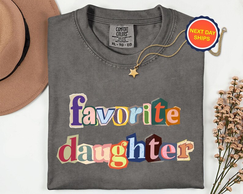 May include: A charcoal grey Comfort Colors t-shirt with the words "favorite daughter" in colourful, cut-out style letters. A gold star pendant necklace and a "Next Day Ships" sticker are also visible.