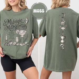 May include: A moss green t-shirt featuring a front graphic with the text Midnight Margaritas, a margarita glass, a frog in a witch hat, a candle, and a bottle. The back graphic displays a vertical arrangement of a moon, a frog, a margarita, a coffee pot, and a mushroom.