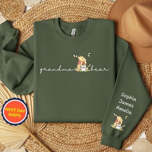 May include: An olive green sweatshirt with the words "grandma bear" in cursive, and a Winnie the Pooh graphic. The right sleeve has the names Sophia, James, and Amelia. A round sticker with the words "NEXT DAY SHIPS" is visible.