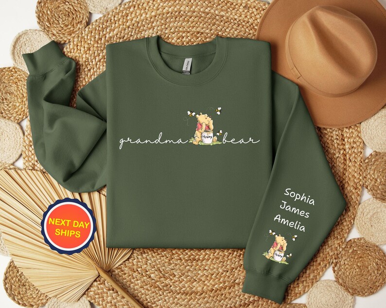 May include: An olive green sweatshirt with the words "grandma bear" in cursive, and a Winnie the Pooh graphic. The right sleeve has the names Sophia, James, and Amelia. A round sticker with the words "NEXT DAY SHIPS" is visible.
