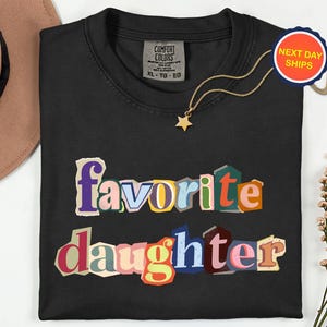 May include: Black crew neck t-shirt with the words "favorite daughter" in a colourful, cut-out style. A gold star pendant necklace and a "Next Day Ships" sticker are visible. The shirt is XL-TG-EG.