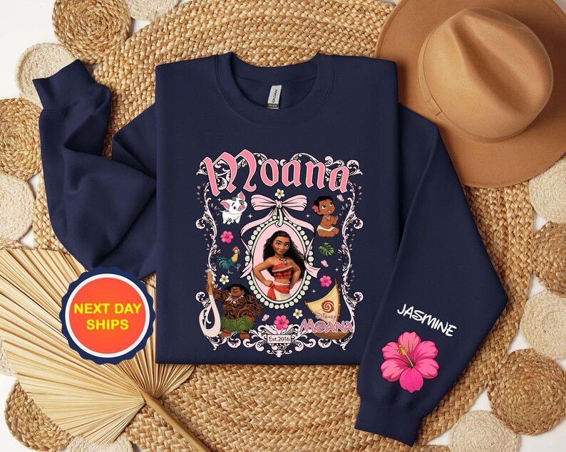Puede incluir: Sudadera azul marino con la palabra "Moana" en escritura rosa sobre una imagen enmarcada del personaje. La manga tiene el nombre "Jasmine" y una flor de hibisco rosa. La sudadera tiene cuello redondo.