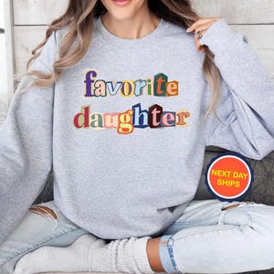 Puede incluir: Una sudadera gris claro con la frase "favorite daughter" en letras de colores. El texto utiliza una variedad de colores, incluyendo azul, naranja y rojo. La sudadera es de estilo casual.