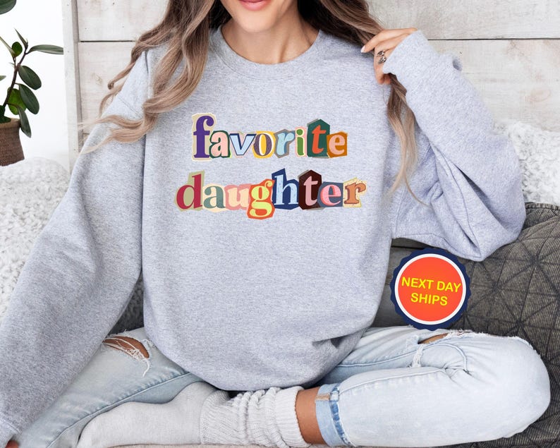 Puede incluir: Una sudadera gris claro con la frase "favorite daughter" en letras de colores. El texto utiliza una variedad de colores, incluyendo azul, naranja y rojo. La sudadera es de estilo casual.