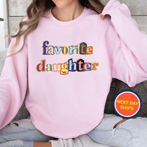 Puede incluir: Sudadera rosa claro con las palabras "favorite daughter" en letras de bloque multicolores. La sudadera tiene mangas largas y cuello redondo. Es visible una pegatina redonda con las palabras "NEXT DAY SHIPS".