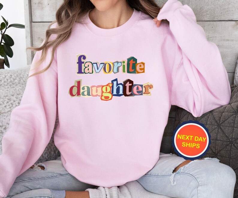 Puede incluir: Sudadera rosa claro con las palabras "favorite daughter" en letras de bloque multicolores. La sudadera tiene mangas largas y cuello redondo. Es visible una pegatina redonda con las palabras "NEXT DAY SHIPS".