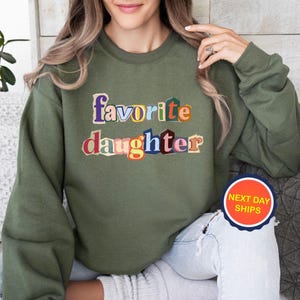 Puede incluir: Sudadera verde oliva con las palabras "favorite daughter" en letras coloridas y superpuestas. El texto es el punto focal de la prenda. La sudadera es de estilo casual.