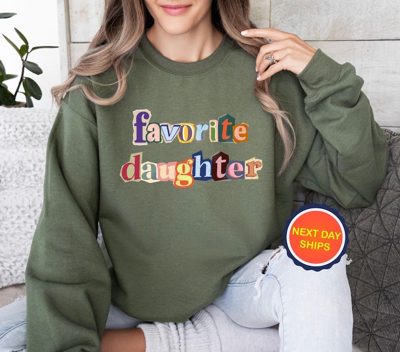 Puede incluir: Sudadera verde oliva con las palabras "favorite daughter" en letras coloridas y superpuestas. El texto es el punto focal de la prenda. La sudadera es de estilo casual.