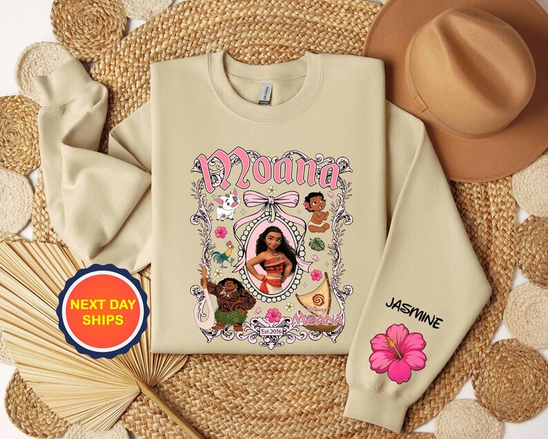 Puede incluir: Sudadera beige con un gr&aacute;fico rosa y blanco de "Moana" e ilustraciones de personajes. La manga derecha muestra el nombre "Jasmine" y una flor de hibisco rosa. Es visible una pegatina de "Next Day Ships".