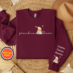 May include: Burgundy sweatshirt with the words "grandma bear" in cursive, and a Winnie the Pooh graphic. The right sleeve has the names Sophia, James, and Amelia. A round sticker with "NEXT DAY SHIPS" is visible.