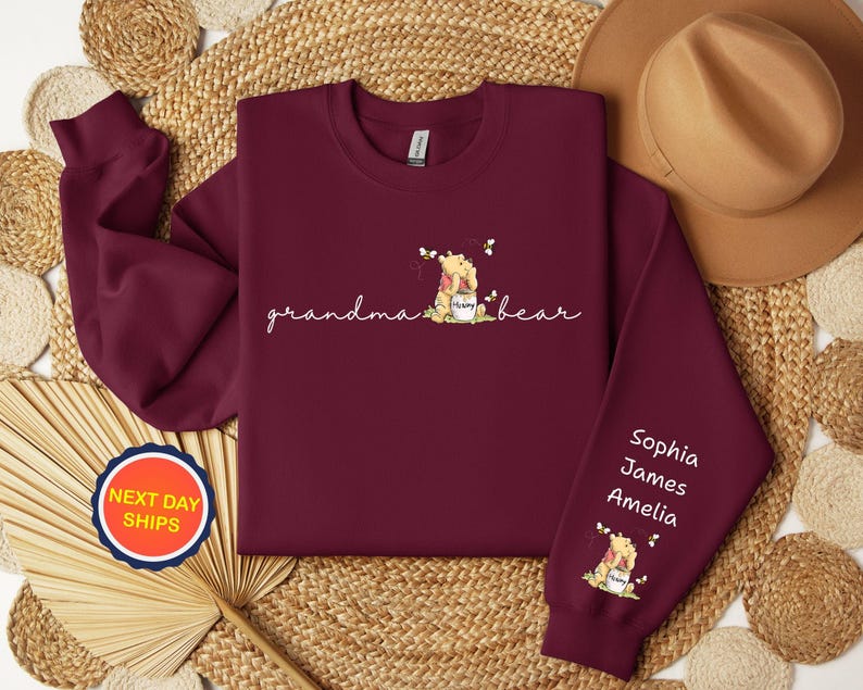 May include: Burgundy sweatshirt with the words "grandma bear" in cursive, and a Winnie the Pooh graphic. The right sleeve has the names Sophia, James, and Amelia. A round sticker with "NEXT DAY SHIPS" is visible.