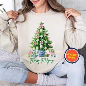 May include: A cream-colored sweatshirt featuring a Christmas tree decorated with pink bows and square ornaments. The tree is set in a blue and white vase, with wrapped gifts at the base. The text "Merry Mahjong" is printed below the tree.