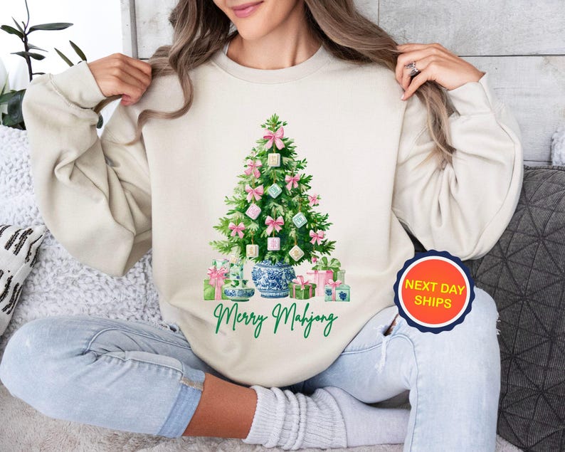 May include: A cream-colored sweatshirt featuring a Christmas tree decorated with pink bows and square ornaments. The tree is set in a blue and white vase, with wrapped gifts at the base. The text "Merry Mahjong" is printed below the tree.
