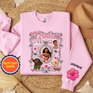 Puede incluir: Una sudadera rosa claro con un dise&ntilde;o de Moana. El dise&ntilde;o incluye el nombre "Moana" en escritura rosa, ilustraciones de personajes y el nombre "Jasmine" en la manga con una flor de hibisco rosa.
