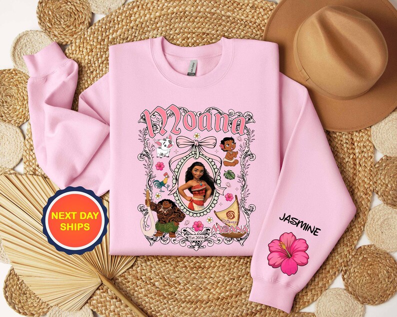 Puede incluir: Una sudadera rosa claro con un dise&ntilde;o de Moana. El dise&ntilde;o incluye el nombre "Moana" en escritura rosa, ilustraciones de personajes y el nombre "Jasmine" en la manga con una flor de hibisco rosa.