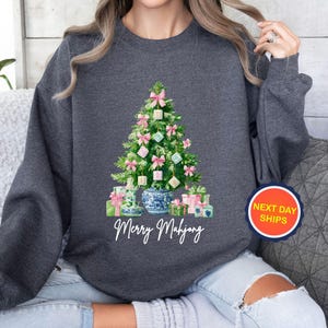 May include: Dark gray sweatshirt featuring a Christmas tree decorated with pink bows and Mahjong tiles. The tree is surrounded by wrapped gifts and a blue and white vase. The text "Merry Mahjong" is below the tree.