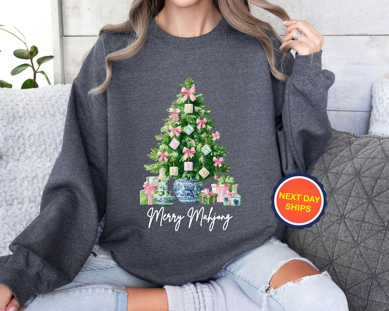 May include: Dark gray sweatshirt featuring a Christmas tree decorated with pink bows and Mahjong tiles. The tree is surrounded by wrapped gifts and a blue and white vase. The text "Merry Mahjong" is below the tree.