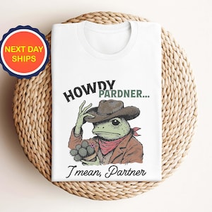May include: White t-shirt featuring a cartoon frog wearing a cowboy hat and holding a cactus. The text "Howdy Pardner... I mean, Partner" is printed on the shirt.