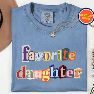 May include: A light blue Comfort Colors t-shirt with the words "favorite daughter" in a colourful, cut-out style. A gold star pendant necklace and a "Next Day Ships" tag are also visible.
