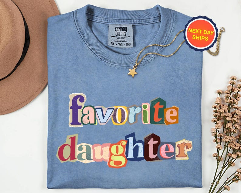 May include: A light blue Comfort Colors t-shirt with the words "favorite daughter" in a colourful, cut-out style. A gold star pendant necklace and a "Next Day Ships" tag are also visible.