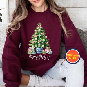 May include: Burgundy sweatshirt featuring a Christmas tree design made of Mahjong tiles, pink bows, and wrapped gifts. The tree sits in a blue and white vase. The text "Merry Mahjong" is below the tree.