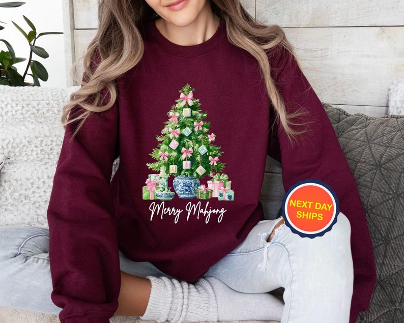 May include: Burgundy sweatshirt featuring a Christmas tree design made of Mahjong tiles, pink bows, and wrapped gifts. The tree sits in a blue and white vase. The text "Merry Mahjong" is below the tree.