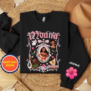 Puede incluir: Sudadera negra con un gr&aacute;fico rosa y blanco del personaje Moana. El dise&ntilde;o incluye la palabra "Moana" en una fuente decorativa, junto con ilustraciones de personajes y flores. La manga tiene el nombre "Jasmine" y una flor de hibisco rosa.