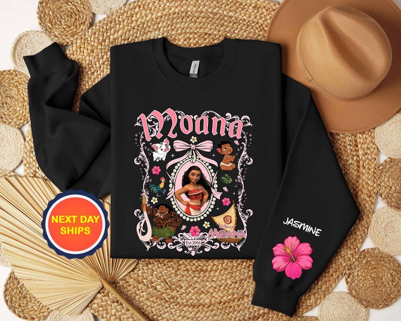 Puede incluir: Sudadera negra con un gr&aacute;fico rosa y blanco del personaje Moana. El dise&ntilde;o incluye la palabra "Moana" en una fuente decorativa, junto con ilustraciones de personajes y flores. La manga tiene el nombre "Jasmine" y una flor de hibisco rosa.