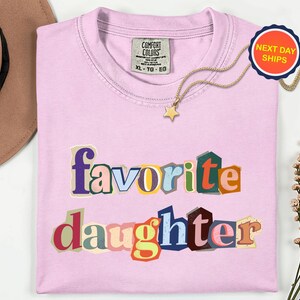 May include: A pale pink t-shirt featuring the words "favorite daughter" in a colourful, cut-out style. A gold necklace with a star pendant is on the shirt. A "Next Day Ships" sticker is visible.